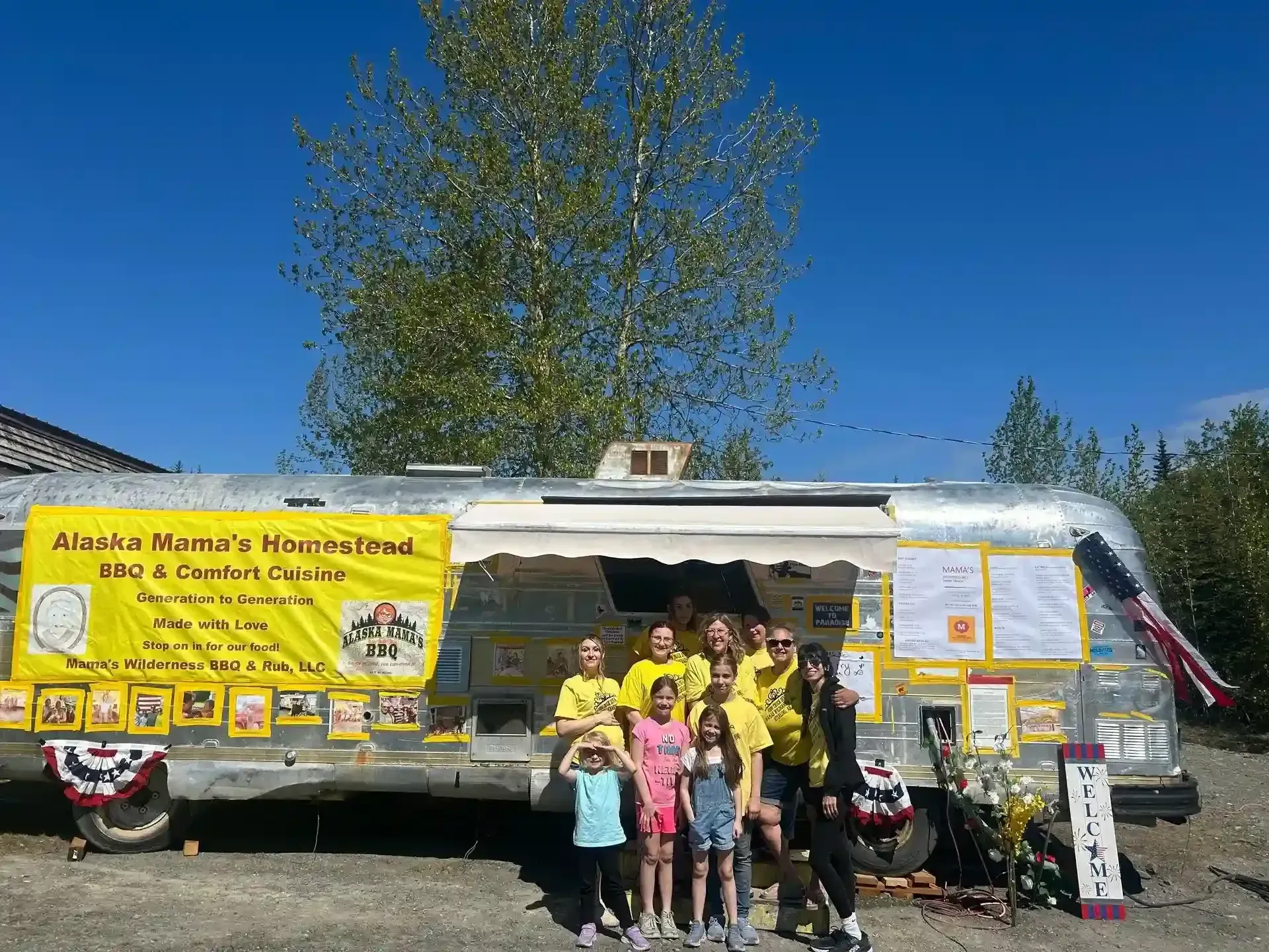 People posing by a yellow food truck at an outdoor event with blue sky and a tree behind it