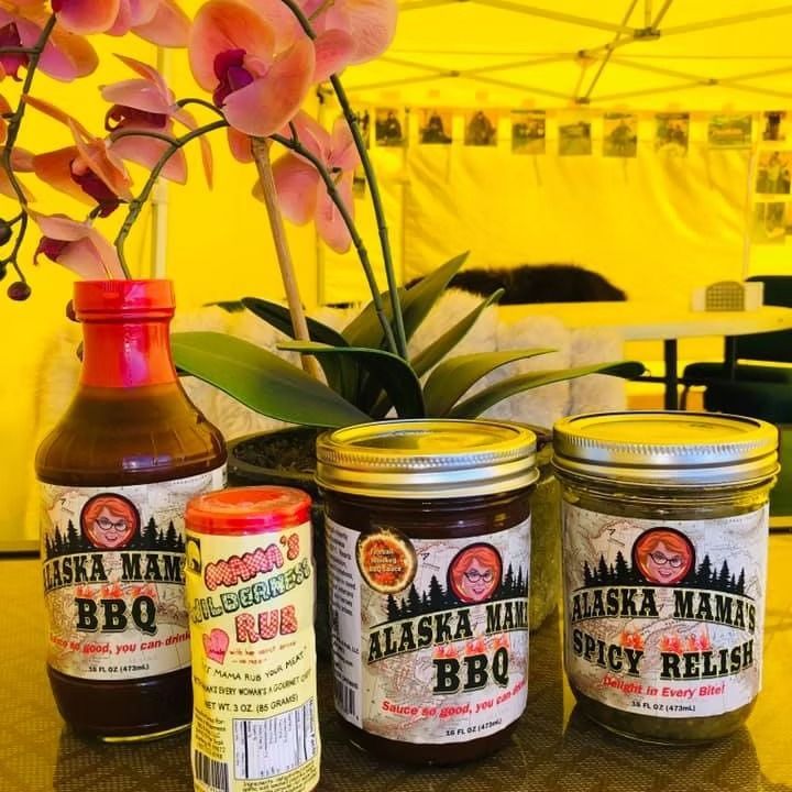 Assorted Alaska Mama BBQ sauces and relish displayed on a table under a yellow tent with orchids behind.