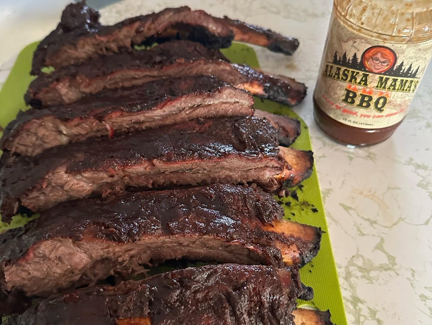 Stack of smoked BBQ ribs on a green tray beside a barbecue sauce bottle