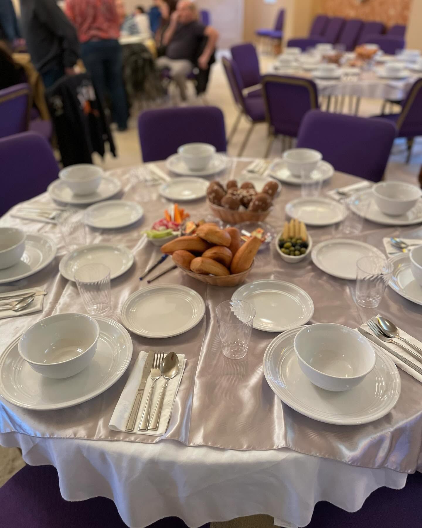 Round banquet table set with white dishes, cups, and a centerpiece of bread and fruit in a purple-carpeted hall