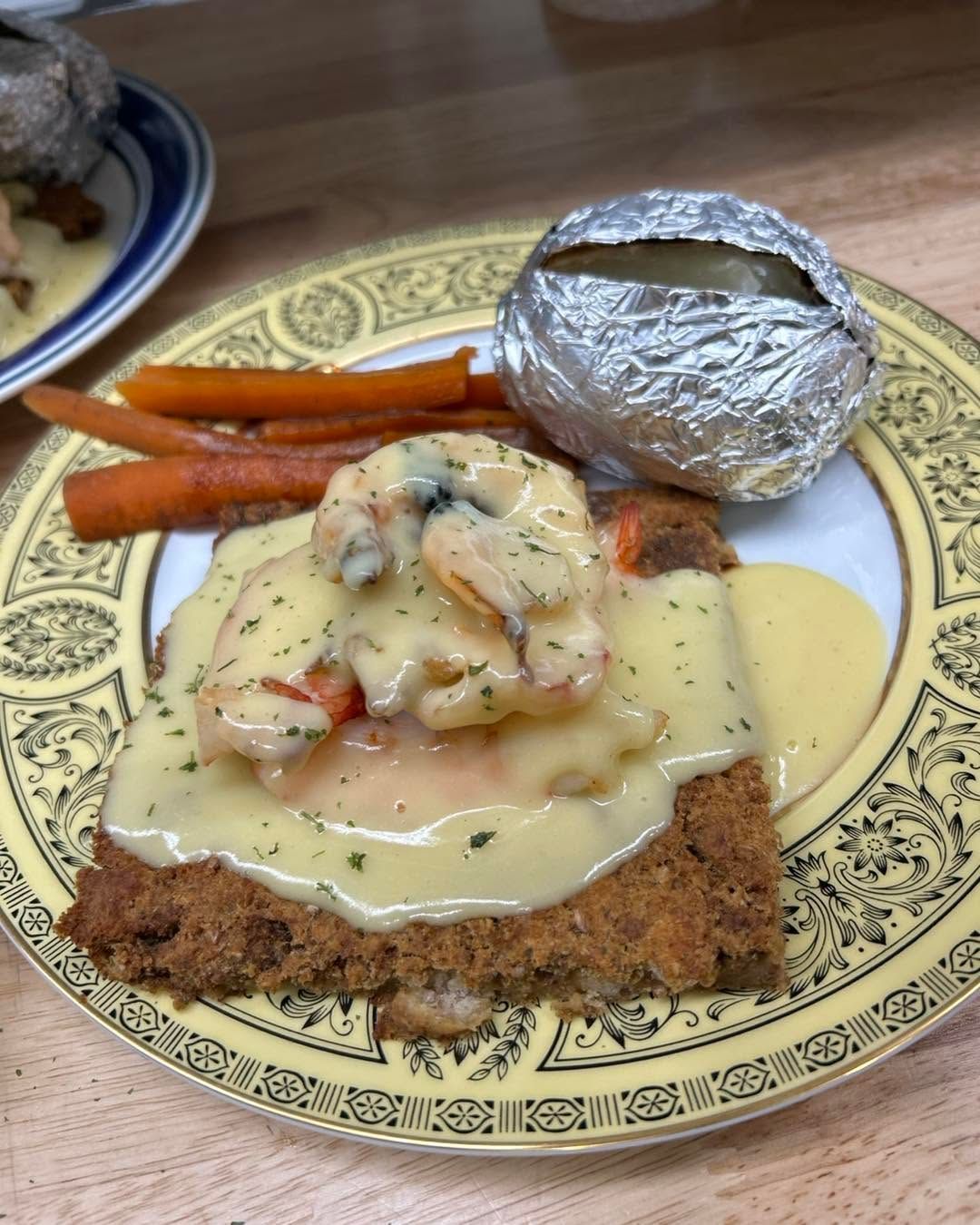 Plate of breaded cutlet with creamy sauce, roasted potatoes, carrots, and foil-wrapped baked potato on a patterned plate