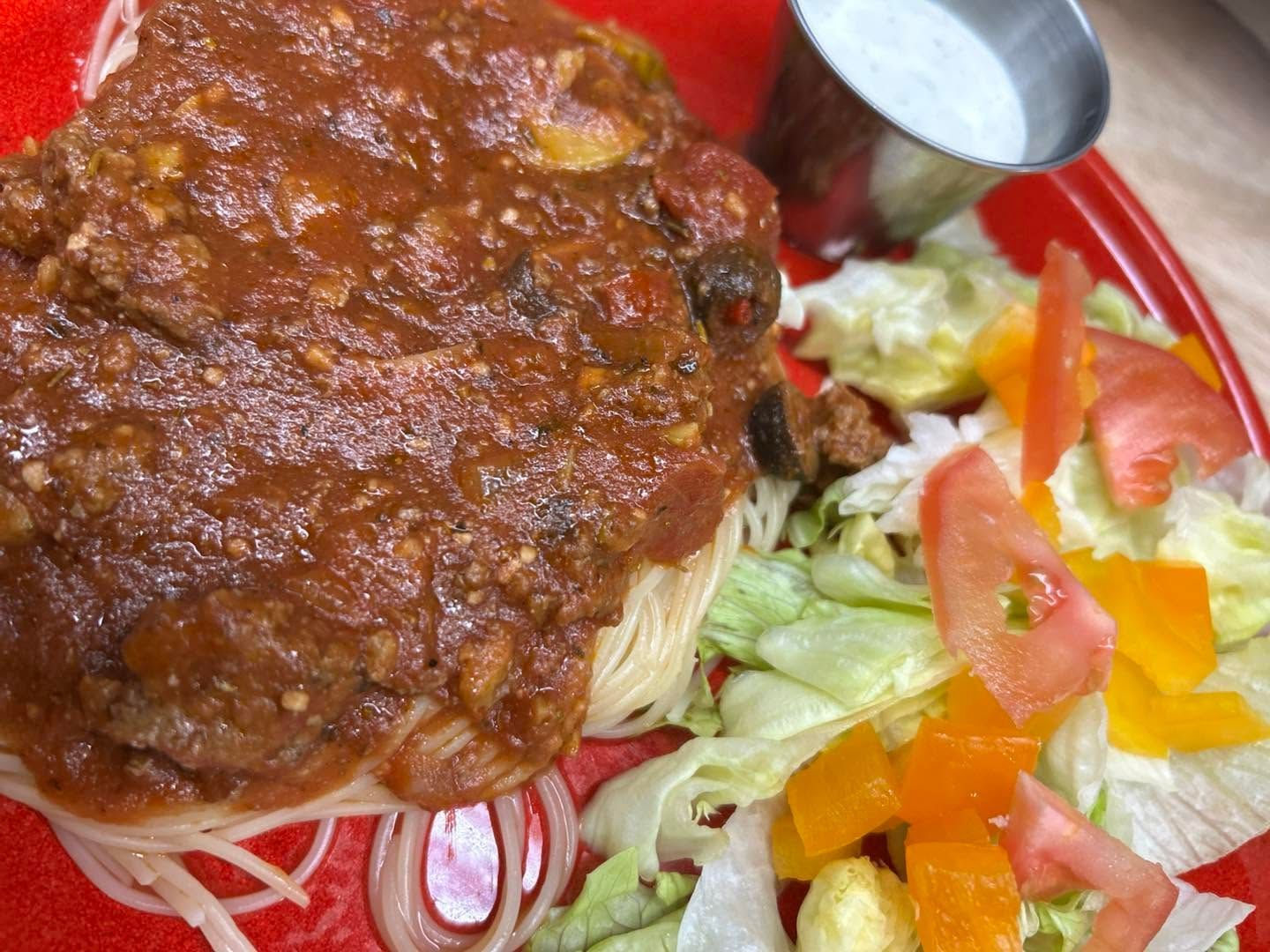 Red plate with spaghetti topped by meat sauce, served beside a fresh salad and a spoon.