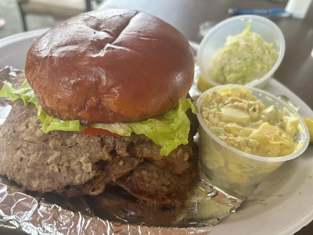 Burger with lettuce on a plate, served with coleslaw and potato salad in small cups.