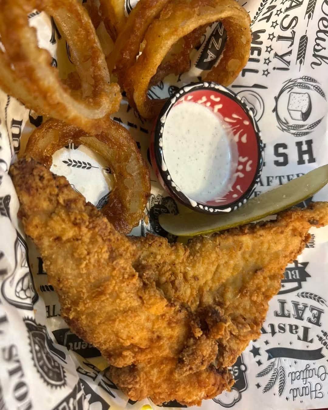 Fried chicken tenders with onion rings and dipping sauce in a basket-lined tray.