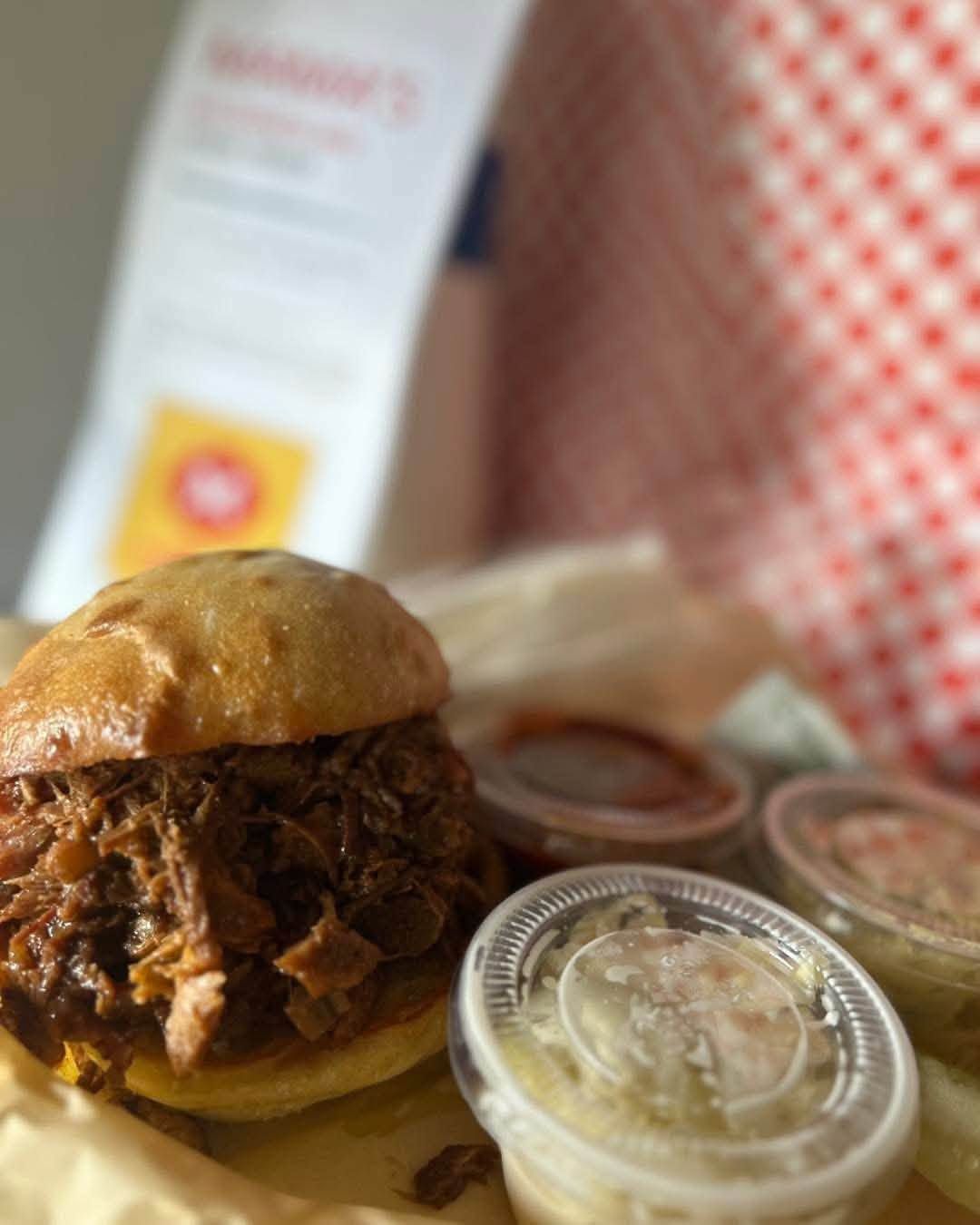 Pulled pork sandwich with dipping sauces on a tray in a red-checkered restaurant setting
