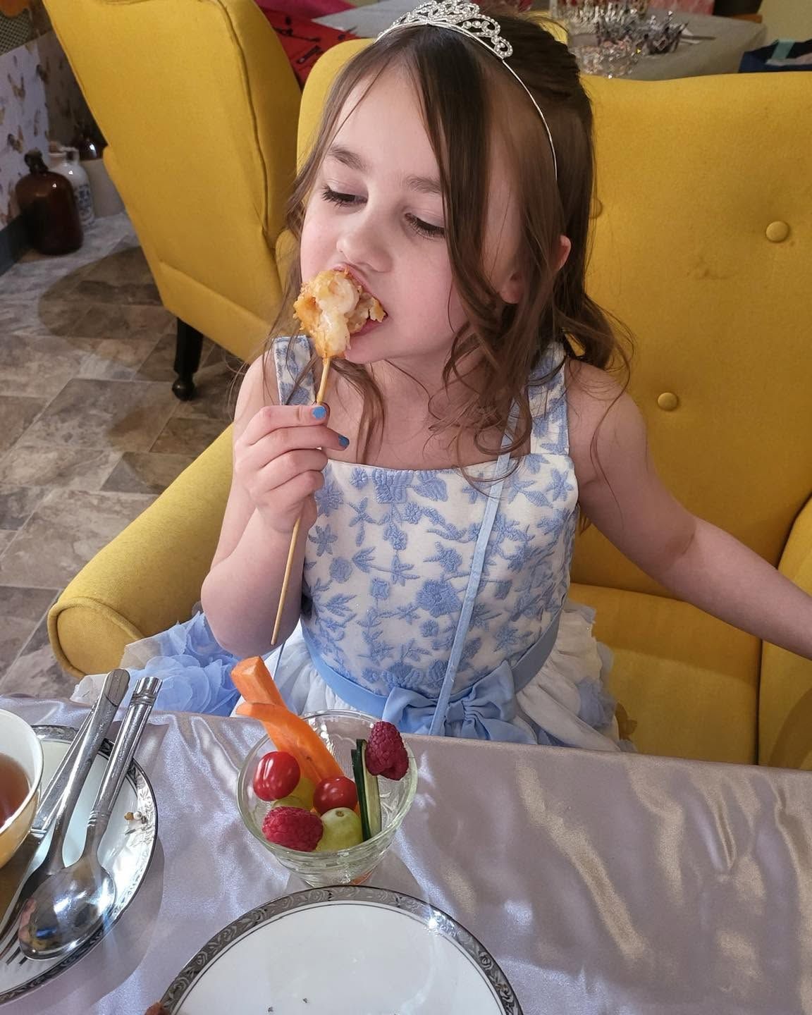 Young girl in a blue dress eating dessert at a table, sitting in a yellow chair.