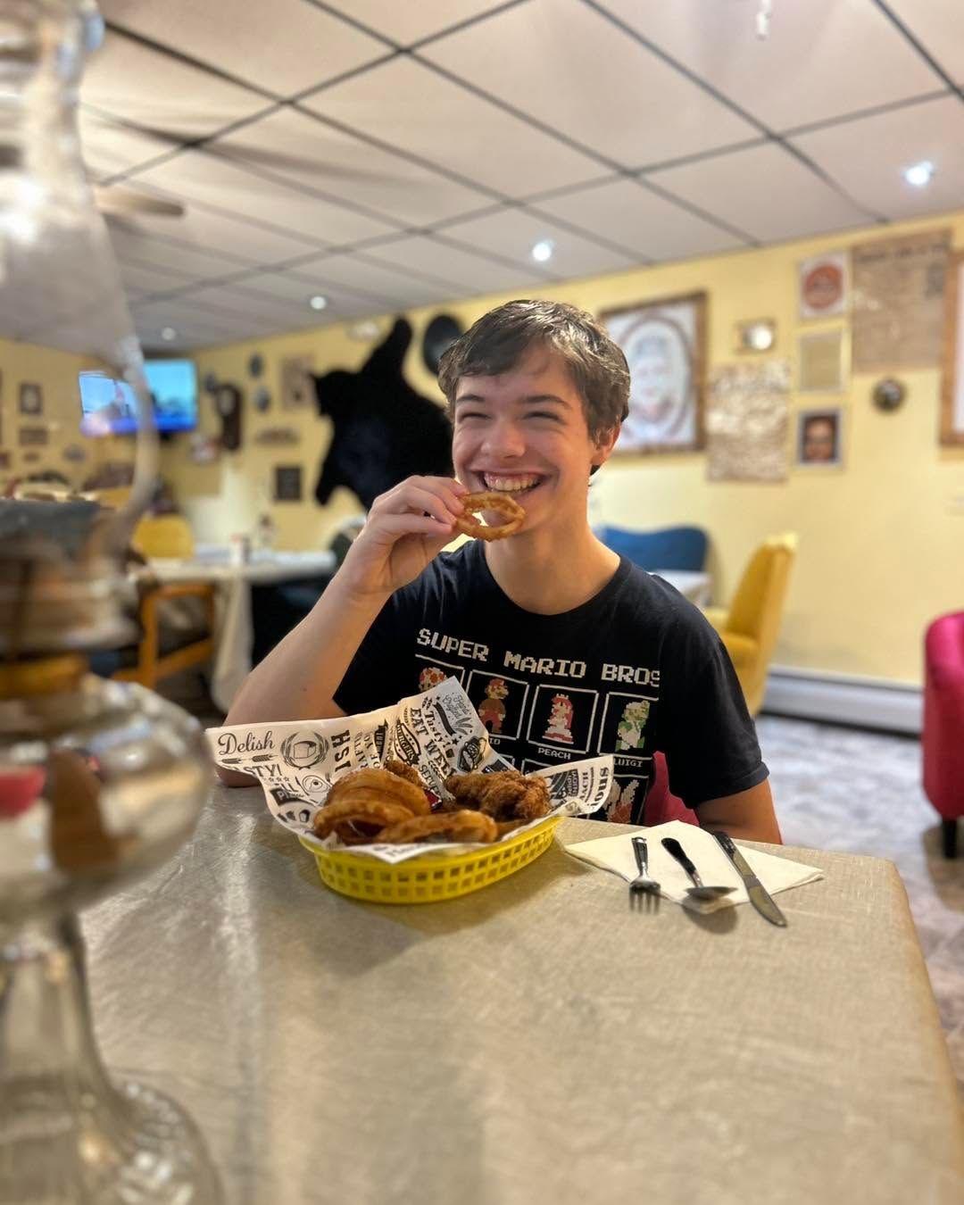 Person smiling while eating fried food at a restaurant table with a basket and utensils.