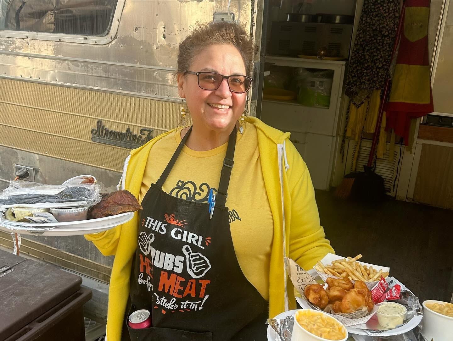 Woman in yellow cardigan and apron smiling, holding trays of food beside a food truck.