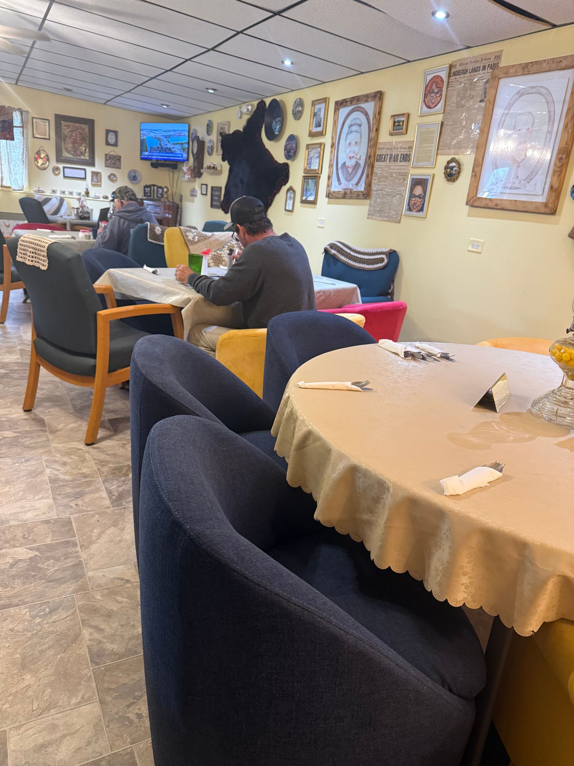 Cozy café lounge with people seated at tables and a TV on the wall, warm beige decor and framed art.