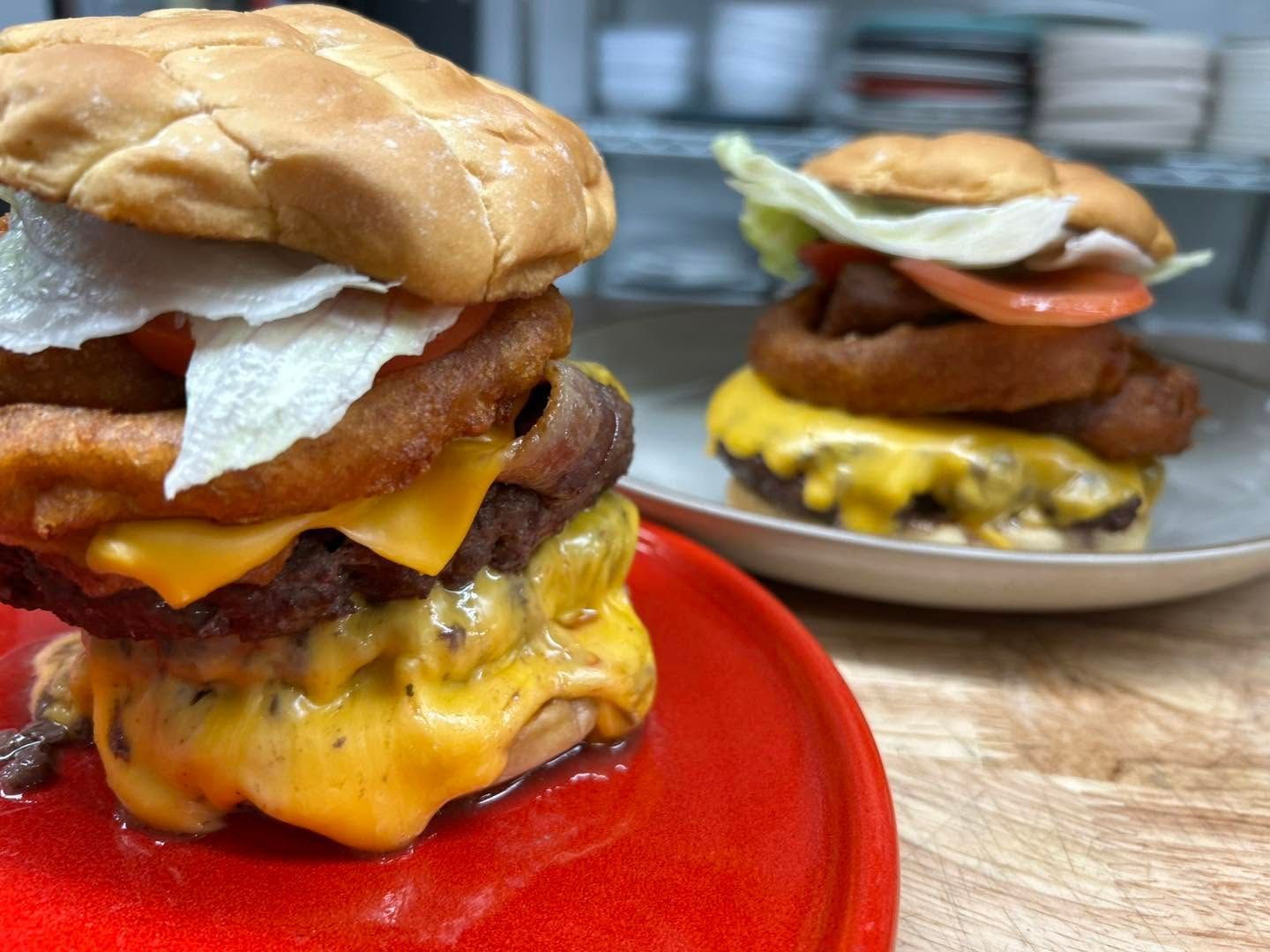 Two stacked cheeseburgers on plates, with melted cheese, bacon, and fried eggs on top.