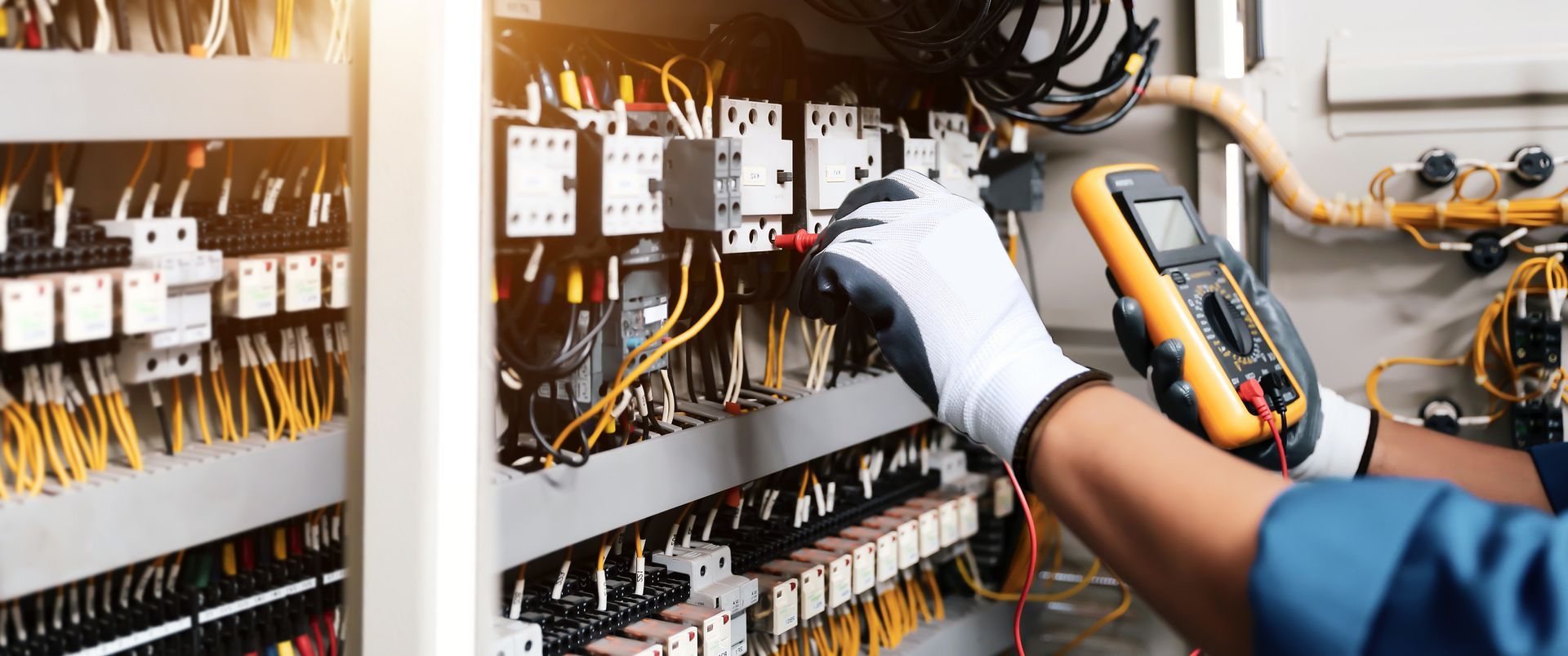 Technician in blue uniform and hard hat wiring an open electrical control panel indoors