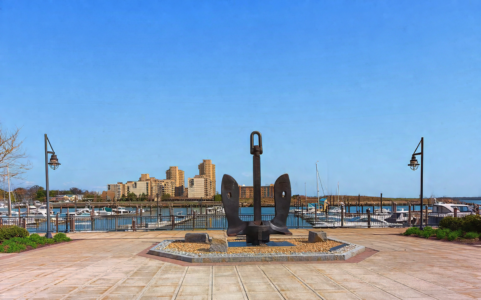 A large iron ship anchor stands on a stone pedestal in a waterfront plaza, overlooking a marina and distant buildings.