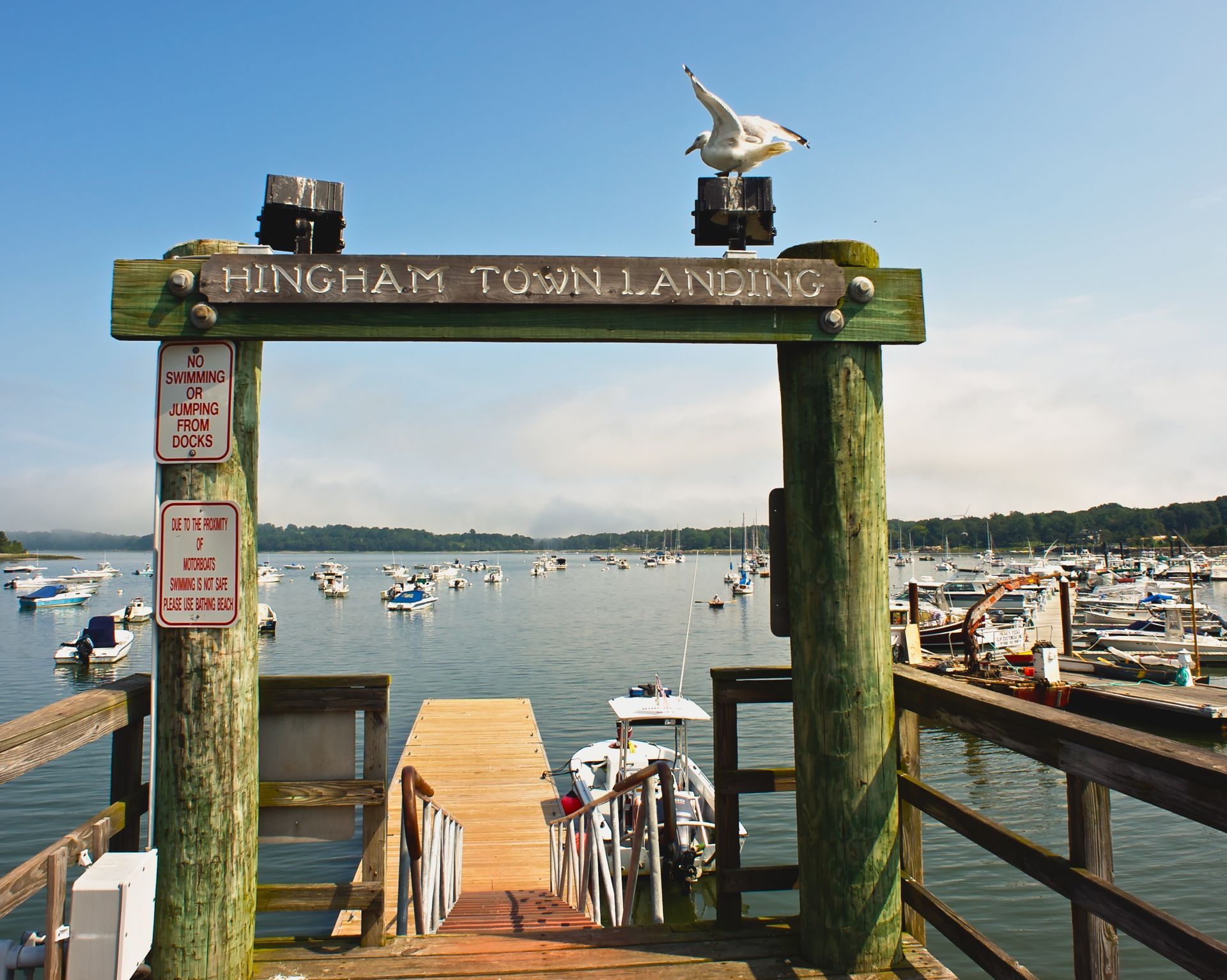 A wooden entrance sign for Hingham Town Landing featuring a perched seagull, overlooking a harbor filled with boats.