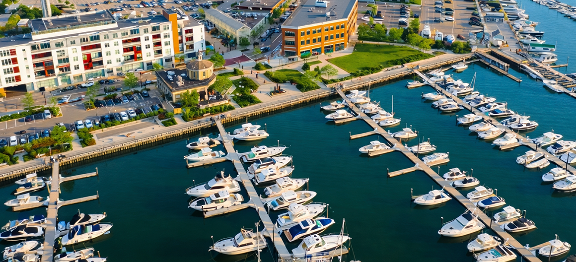 An aerial view of a marina with many boats docked in slips, next to a multi-story building and waterfront pathways.