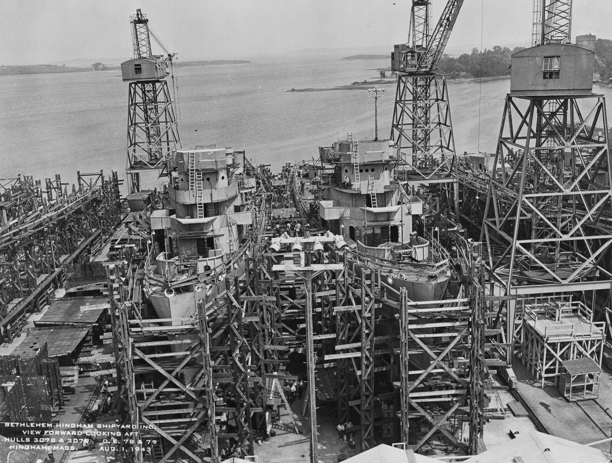 Two naval ships under construction in a shipyard, surrounded by scaffolding and large cranes against a waterfront backdrop.