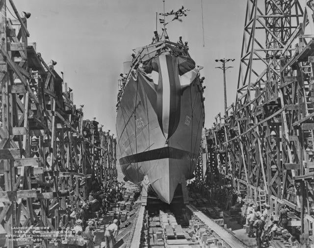 A ship under construction in a dry dock, surrounded by tall wooden scaffolding and workers on the ground.