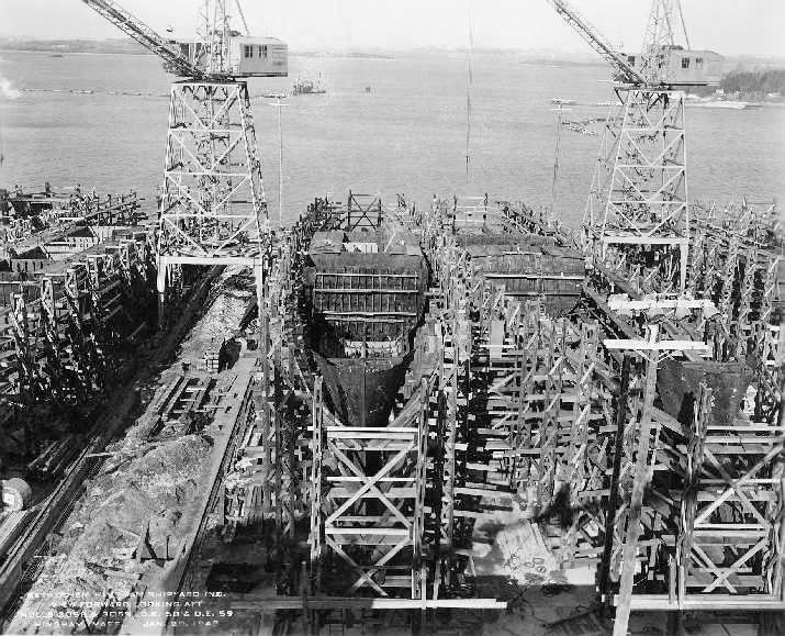 Black and white photograph of a shipyard showing two large cranes towering over a ship under construction.