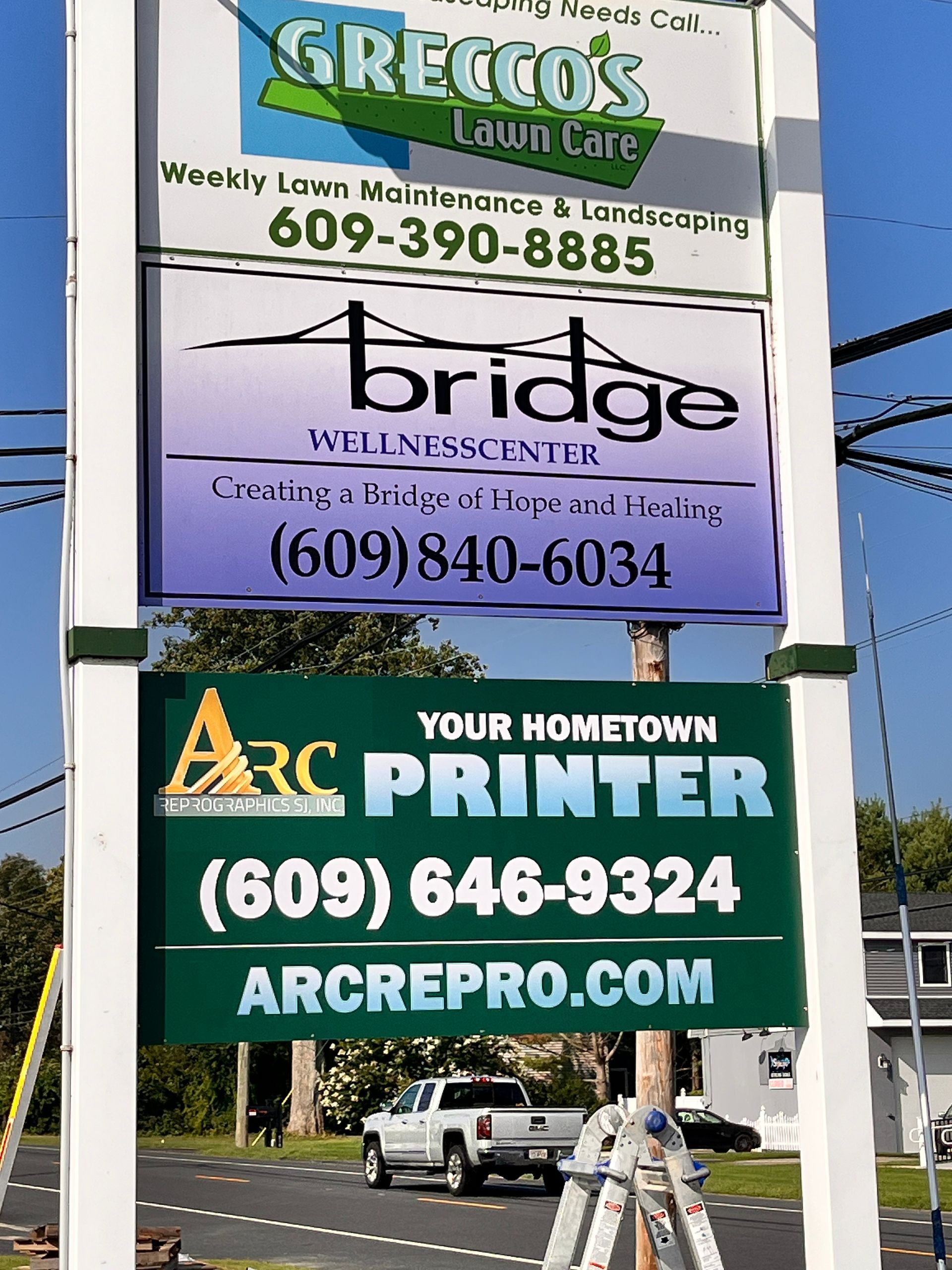 Roadside signboards advertising Grecco’s, Bridge, and Arc Your Hometown Printer with phone numbers