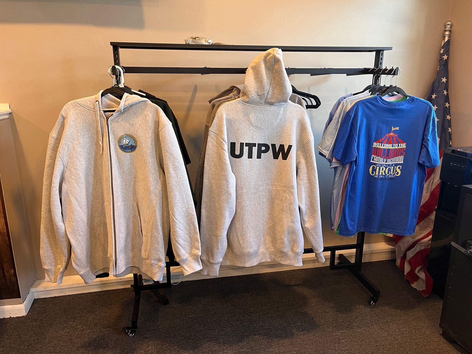 Clothing rack with gray hoodies and blue shirts in a room, with an American flag beside it.