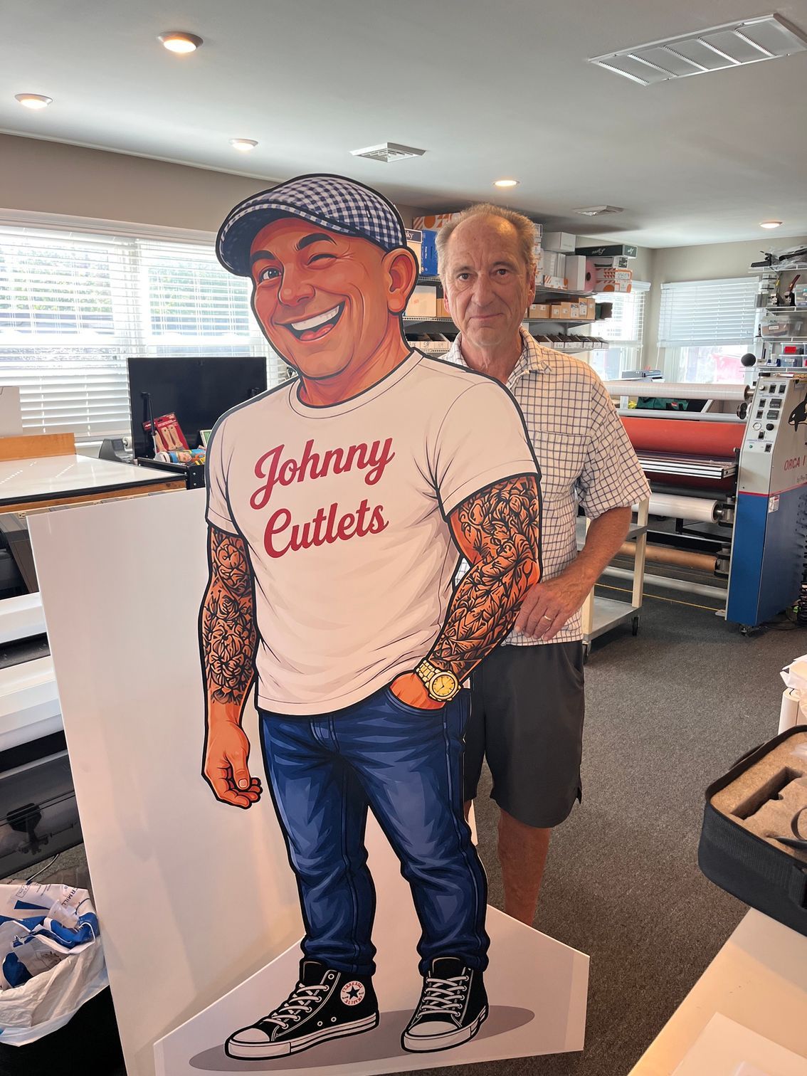 Cardboard cutout of a smiling “Johnny Cutlets” figure in a T-shirt, displayed indoors with a man behind it
