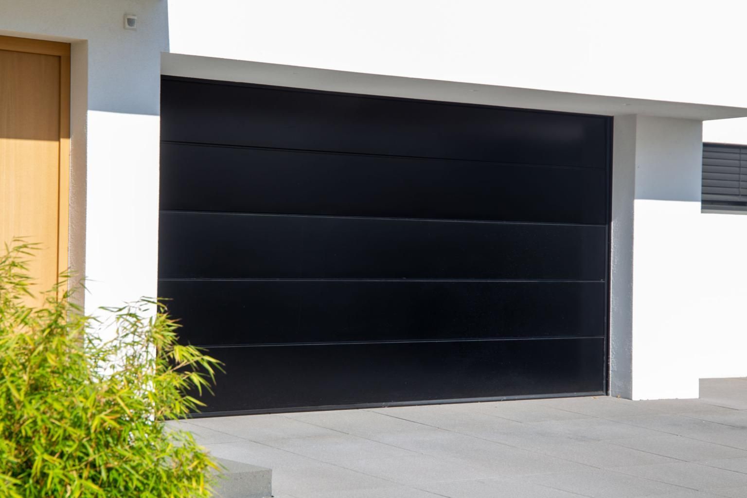 Modern black garage door on a white house with driveway and shrubs