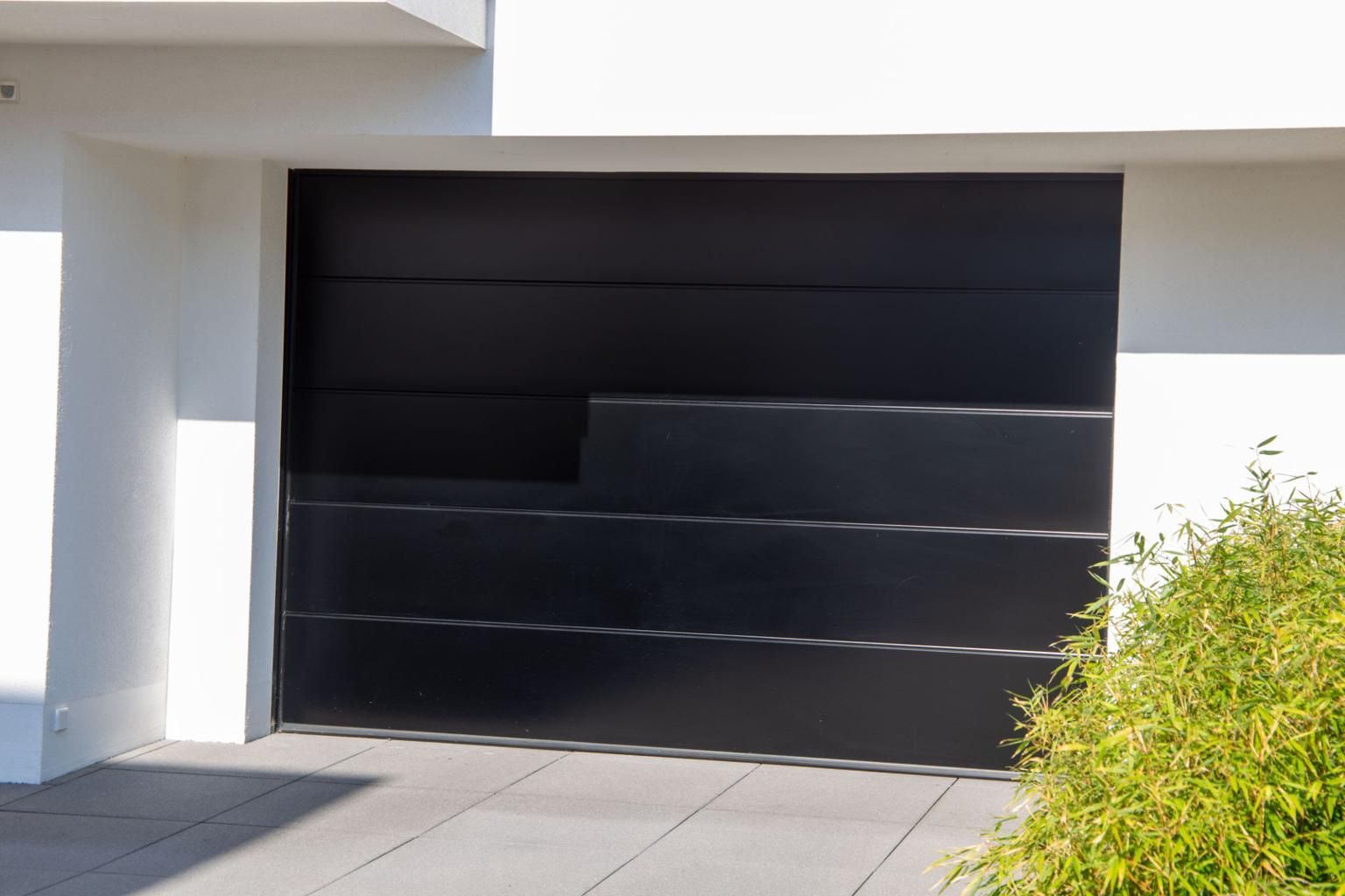 Black modern garage door on a white house exterior with a paved driveway and green shrubbery nearby