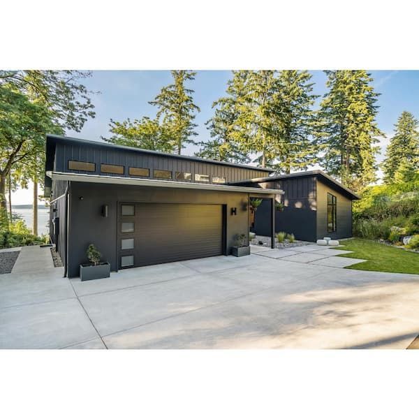 Modern dark-gray house with sloped roofs, large garage door, and driveway surrounded by tall trees