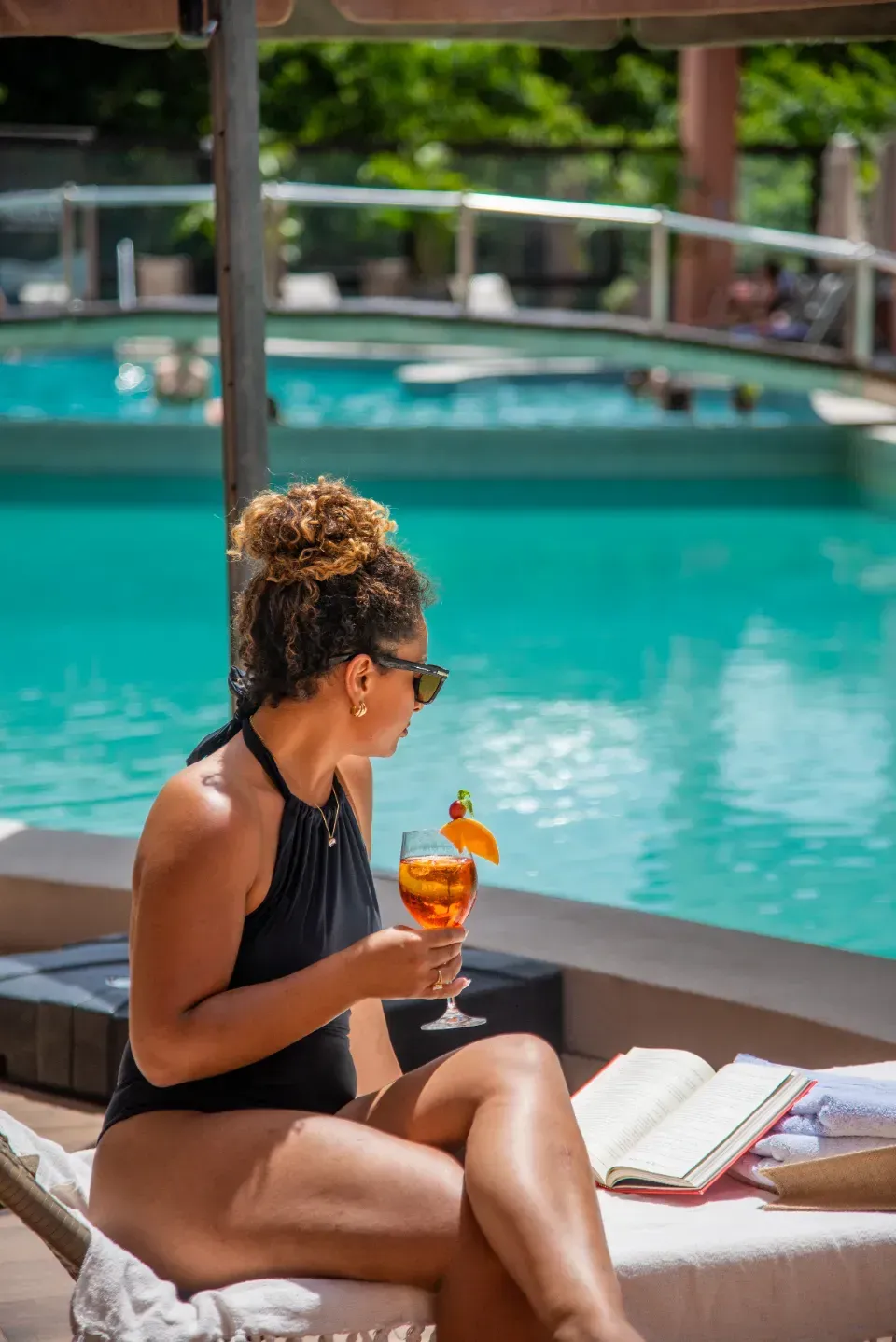 Pessoa relaxando à beira da piscina com uma bebida e um livro em uma espreguiçadeira.
