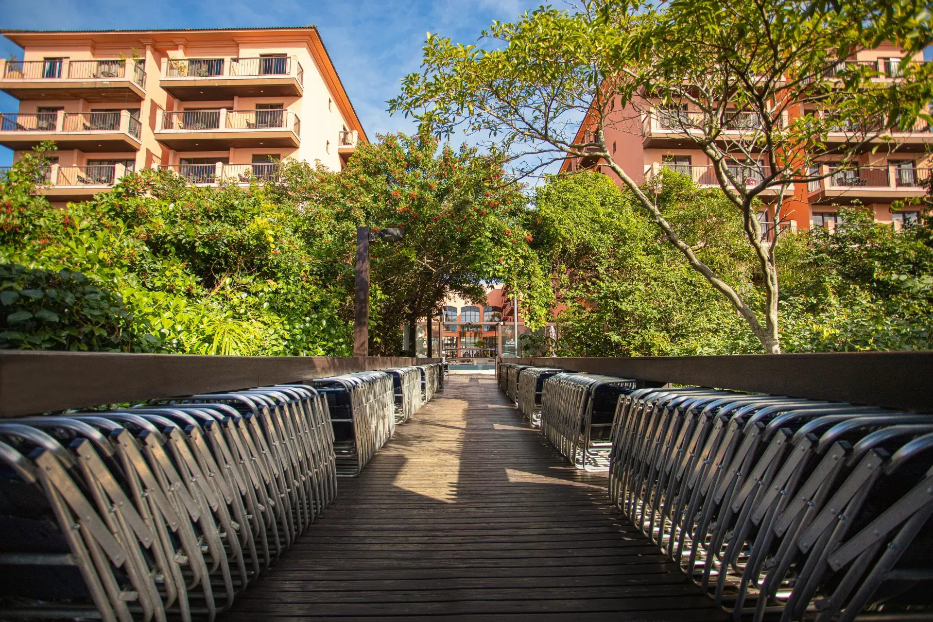Passarela de madeira com corrimão que atravessa um parque verdejante em direção a prédios de apartamentos, banhada de sol.