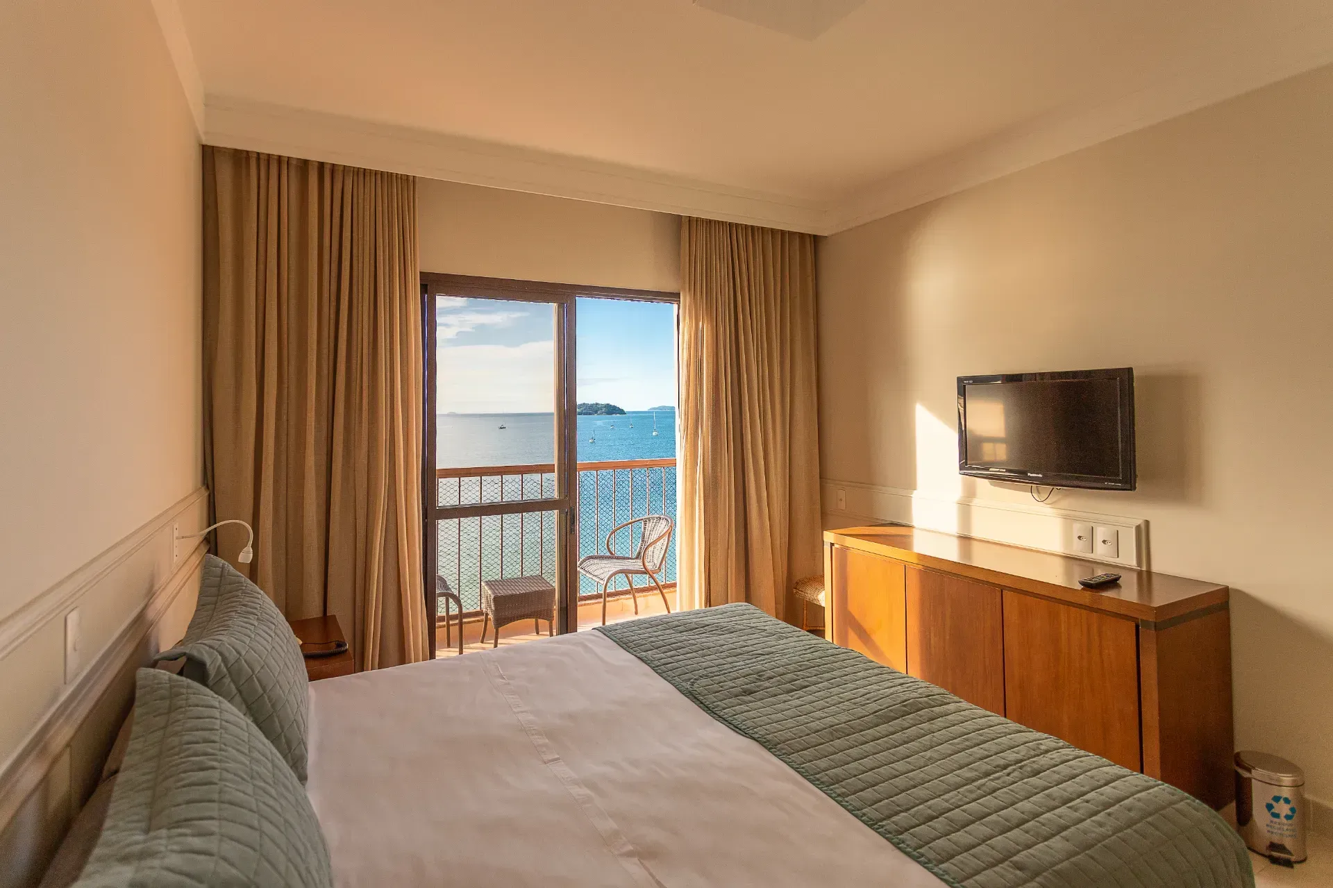 Quarto de hotel com cama, TV e portas de correr que dão para uma varanda com vista para o mar.