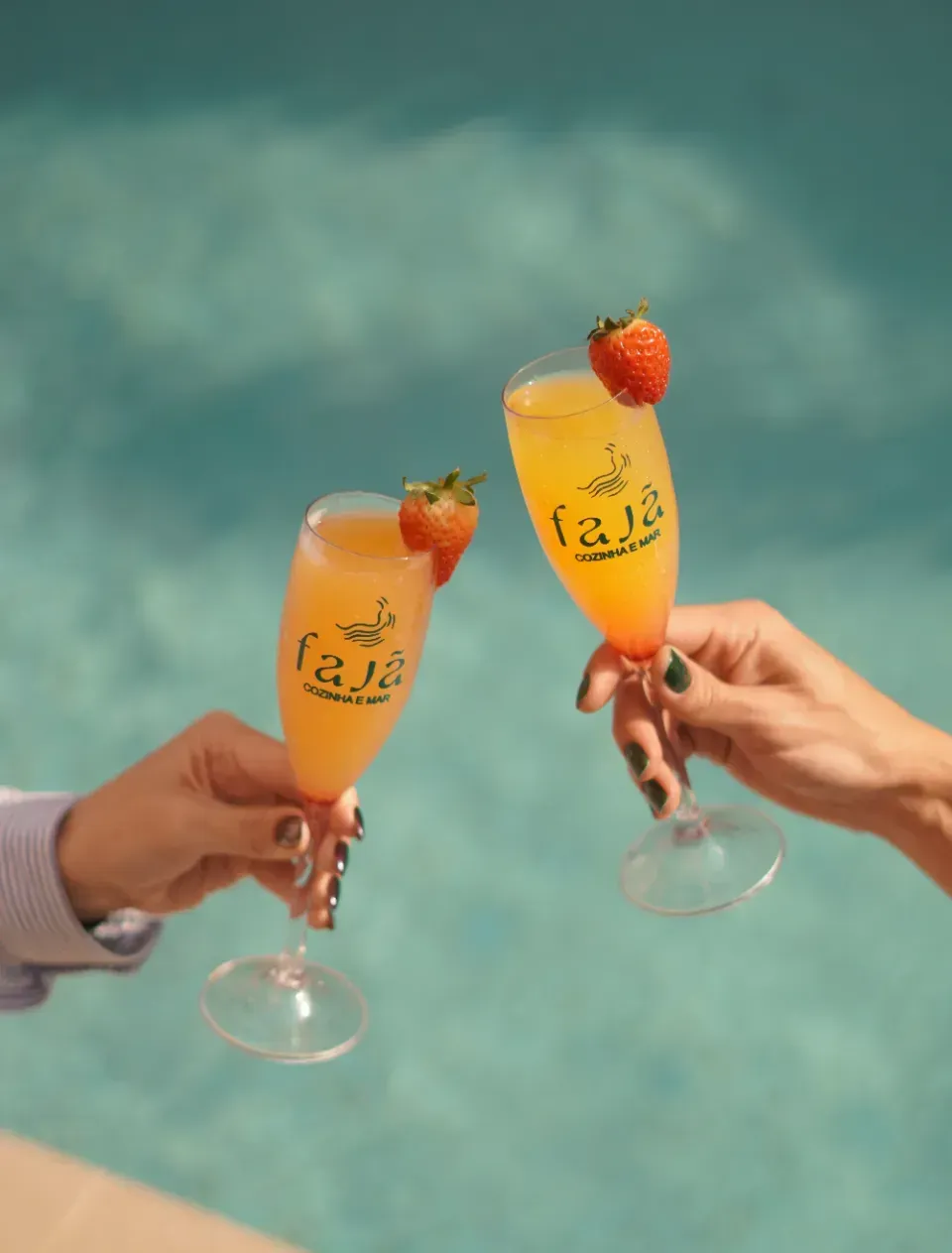 Dois brindes com taças de champanhe laranja decoradas com morangos à beira de uma piscina azul.