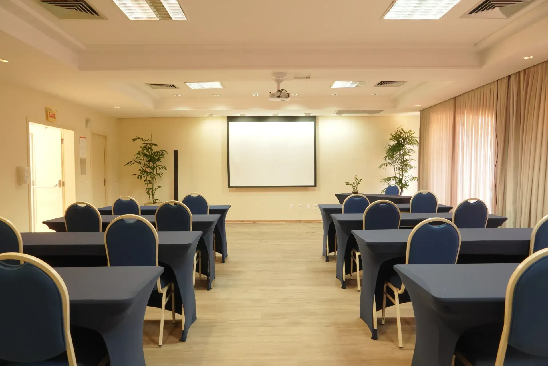 Sala de conferências com fileiras de cadeiras e mesas azuis voltadas para uma tela de projeção sob iluminação aconchegante.