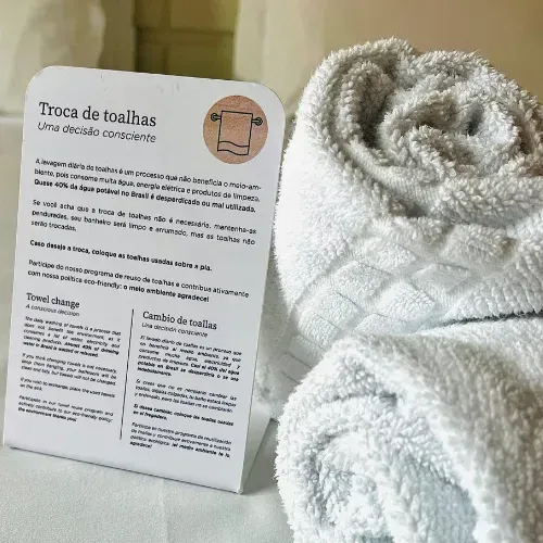 Toalha branca enrolada sobre um cartão, provavelmente de embalagem, ao lado de um tecido branco felpudo sobre um fundo claro.