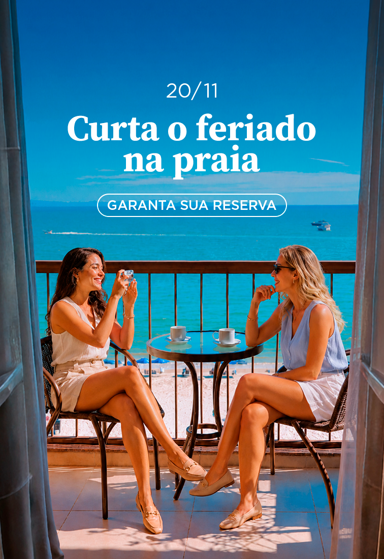 Duas pessoas brindam à mesa na varanda, com vista para o mar; o texto diz: “Aproveite o feriado na praia”.
