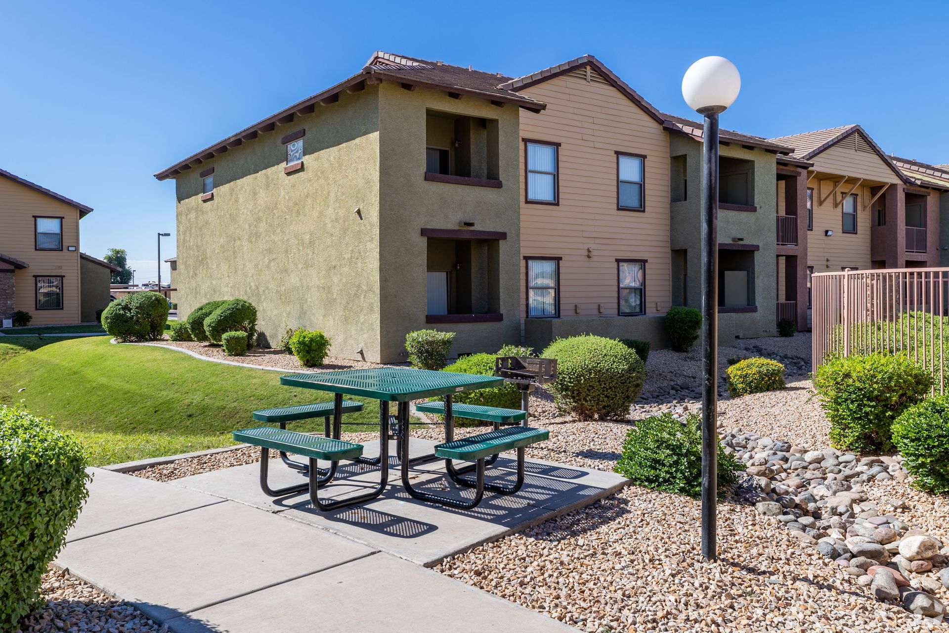 A green picnic table on a concrete patio in front of multi-story apartment buildings with desert landscaping.