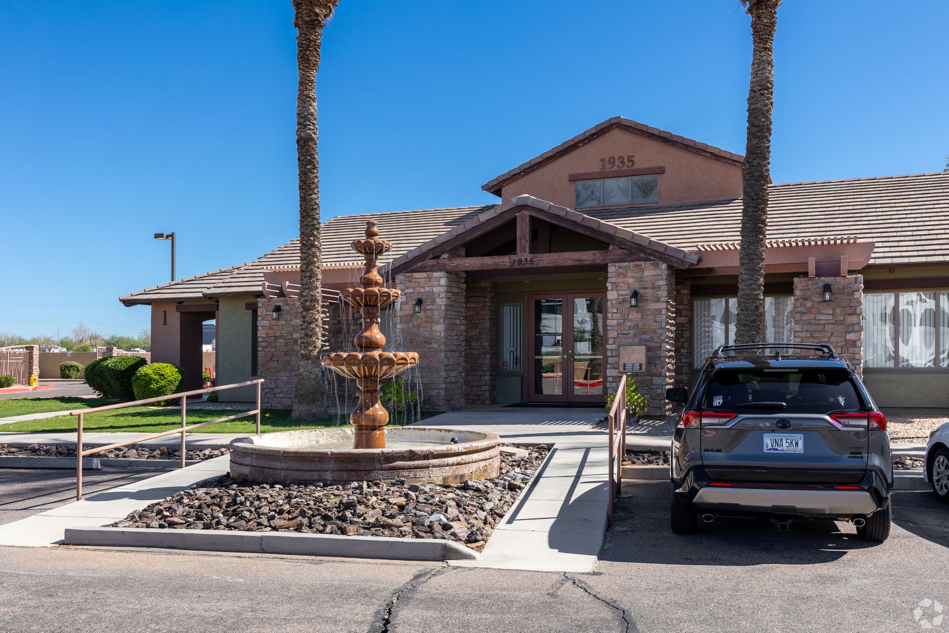 A stone building entrance with a tiered fountain in the foreground, two palm trees, and a parked SUV under a blue sky.