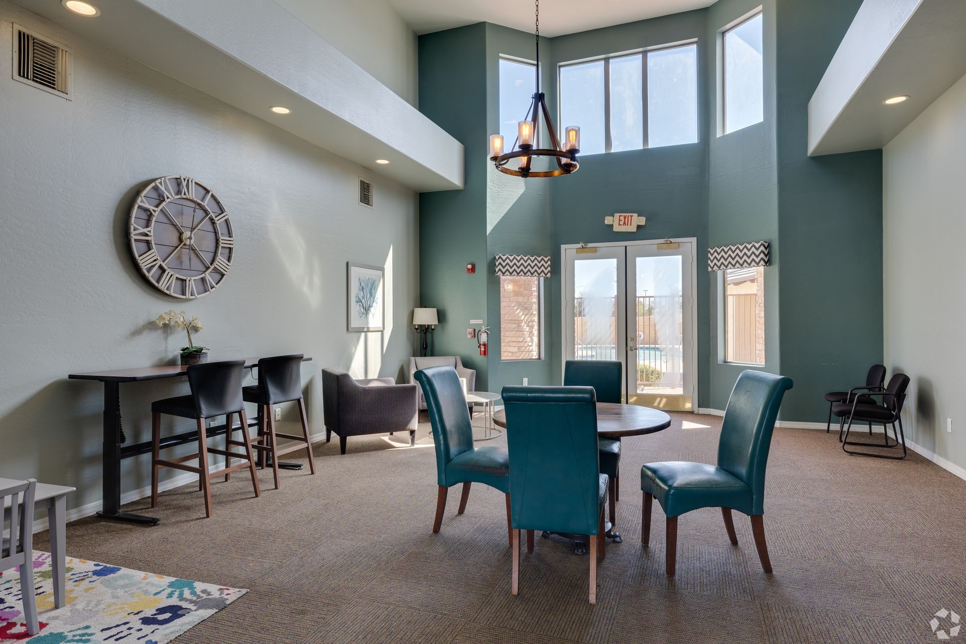 A spacious lobby featuring a high-ceiling lounge with teal walls, a modern chandelier, cafe seating, and a large wall clock.