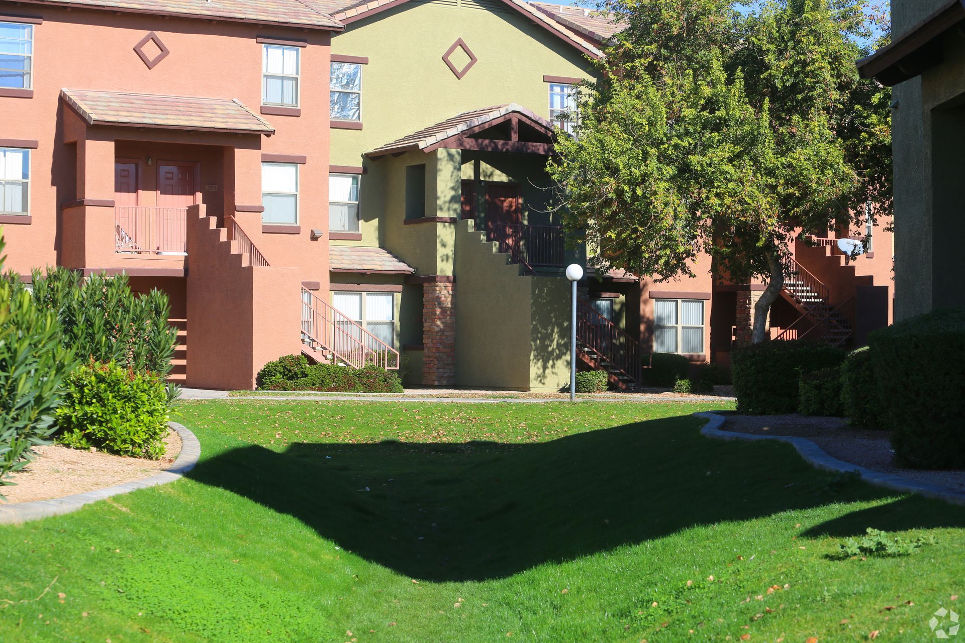 An exterior view of a multi-story apartment complex with stucco walls, a green lawn, and stairs leading to balconies.