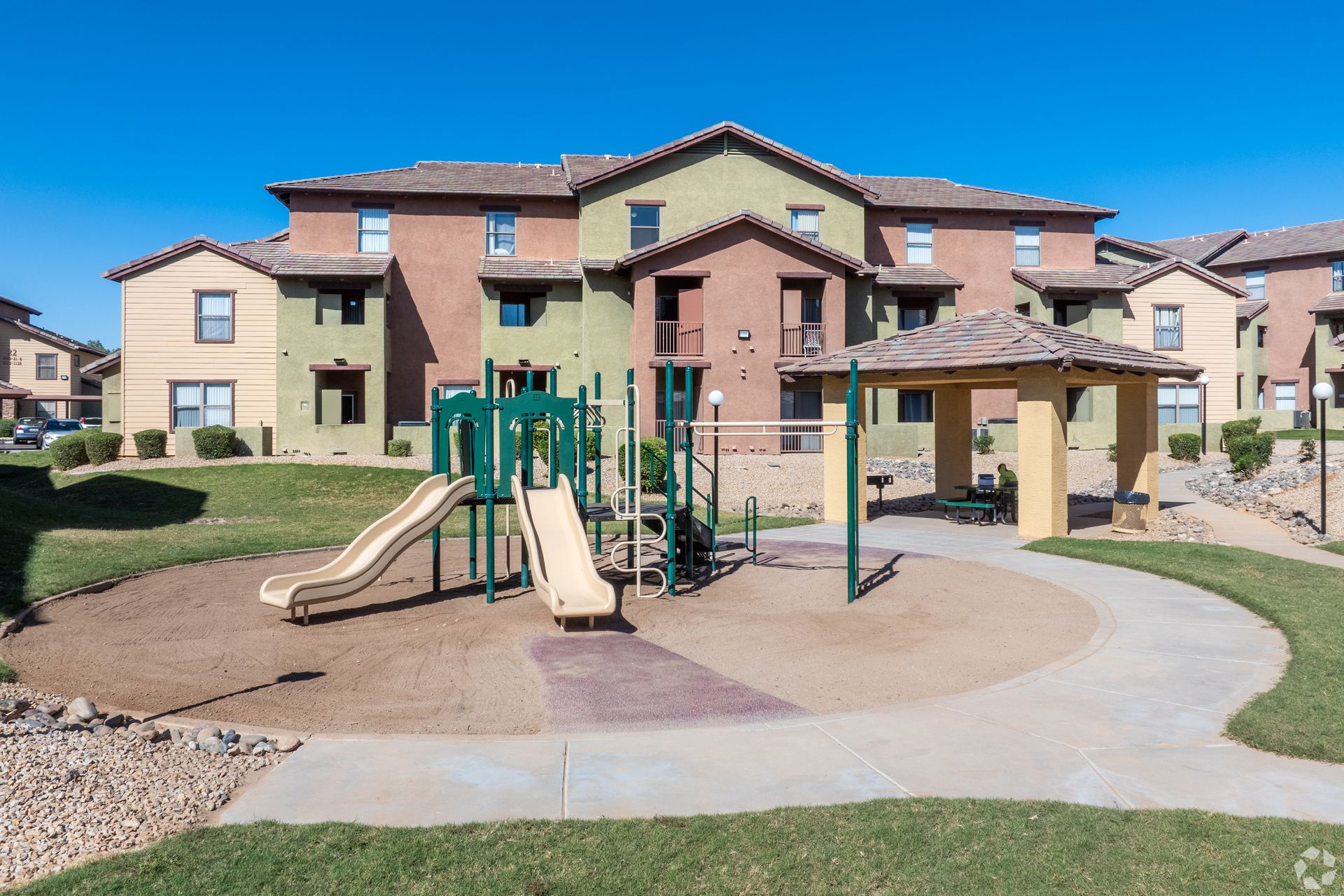 A playground with slides and a covered gazebo sits in front of multi-story, earth-toned apartment buildings under blue sky.