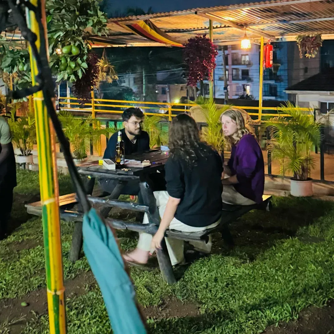 Three people sit at an outdoor picnic table at night in a lit garden café.