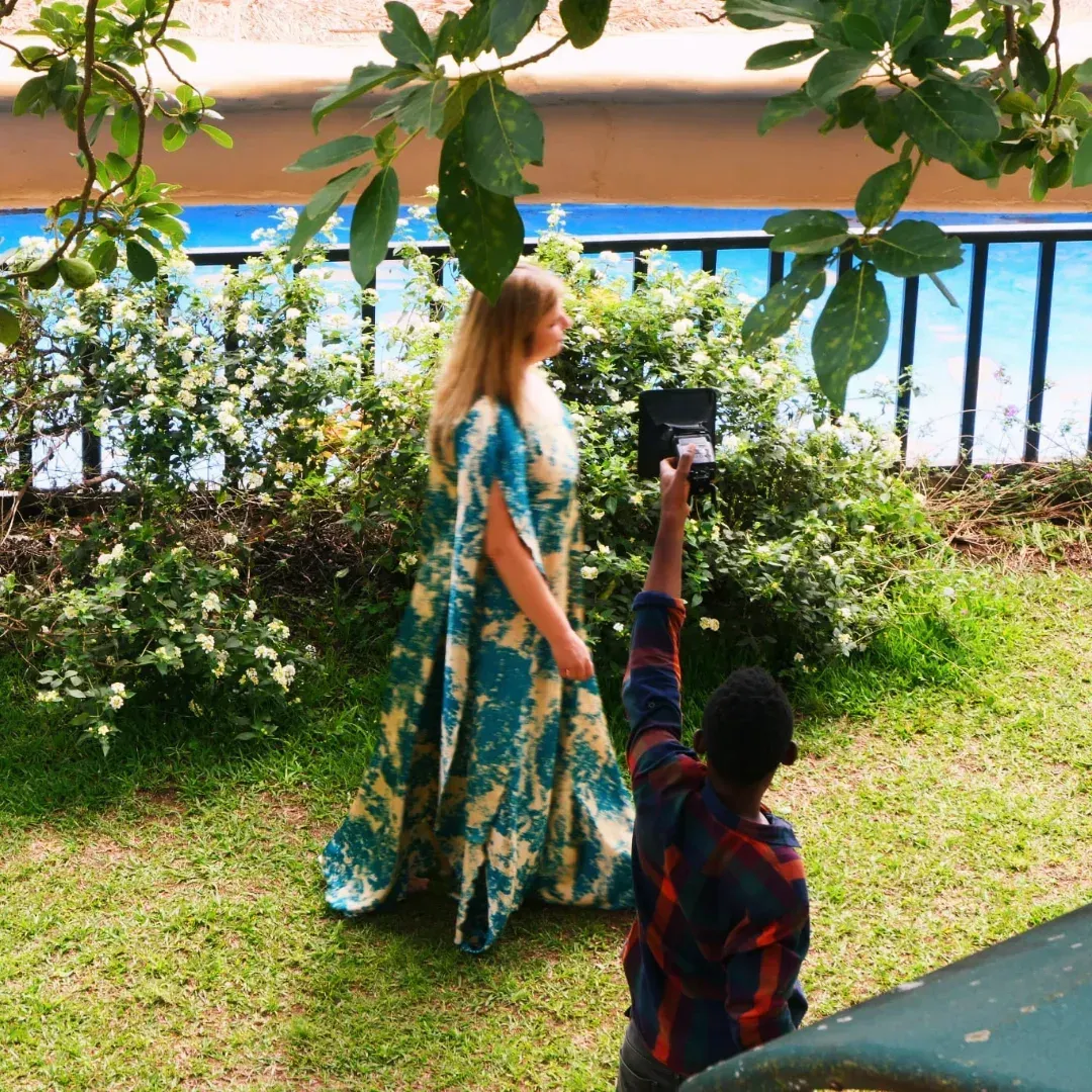Woman in a floral dress walks by a waterfront while a child holds up a phone to take her photo.