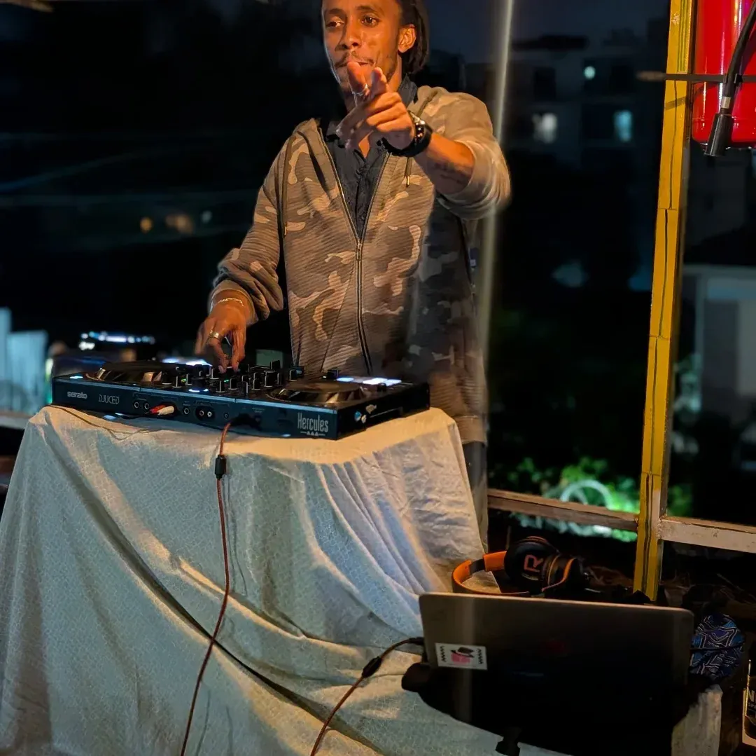 DJ performing at a mixer under warm lights, pointing toward the camera beside a draped table and laptop