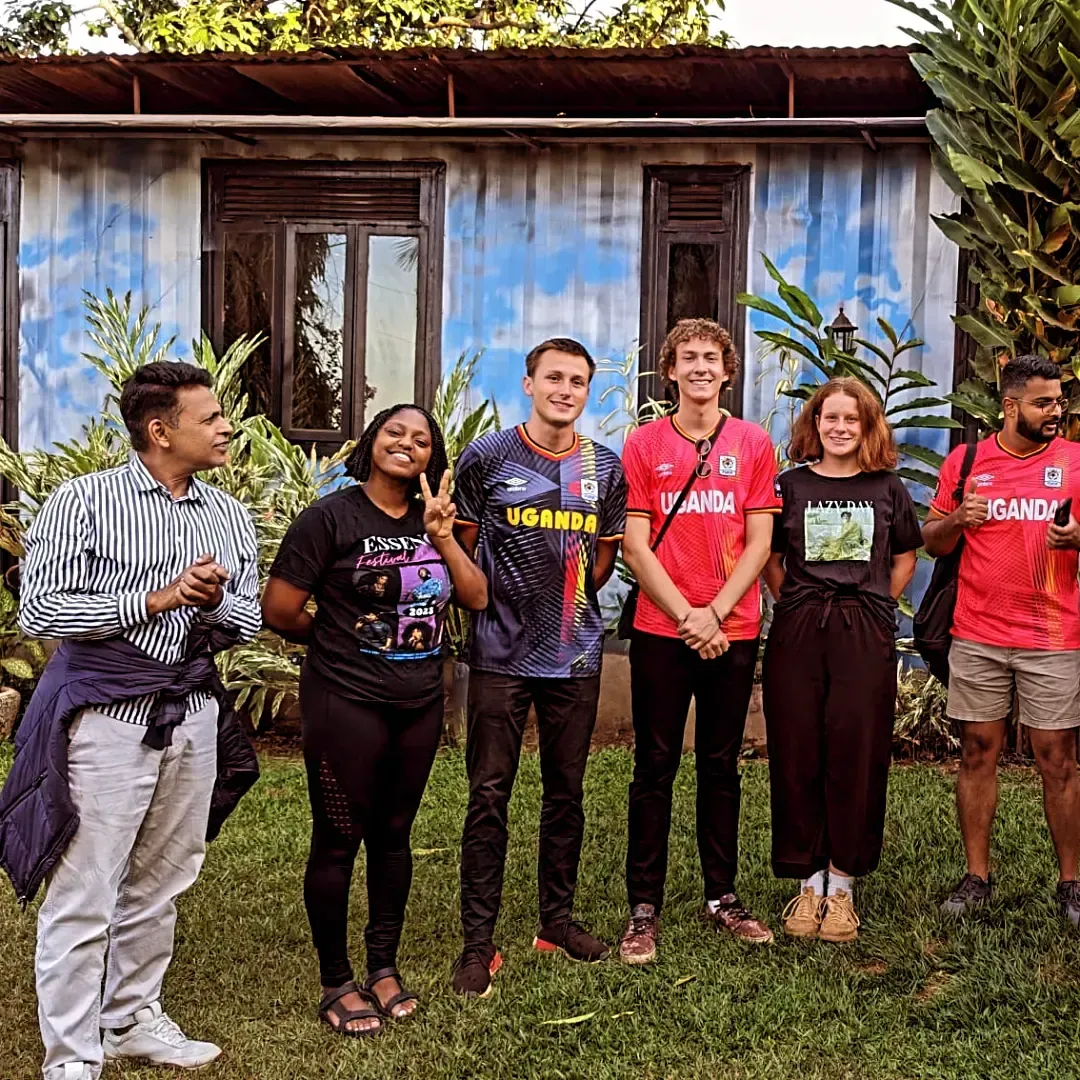 Six people stand smiling in front of a weathered blue house with plants and grass around them.