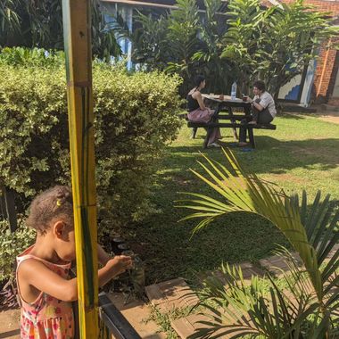 Child peeking from behind a post in a sunny garden with people sitting at a picnic table in the background