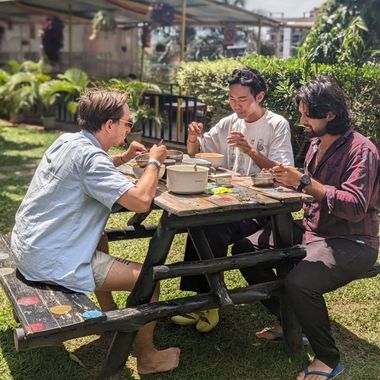 Three people eating together at a picnic table in a sunny outdoor garden.