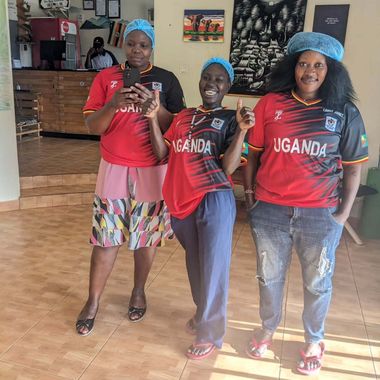 Three people in Uganda jerseys and blue hairnets posing indoors, smiling and holding up drinks.