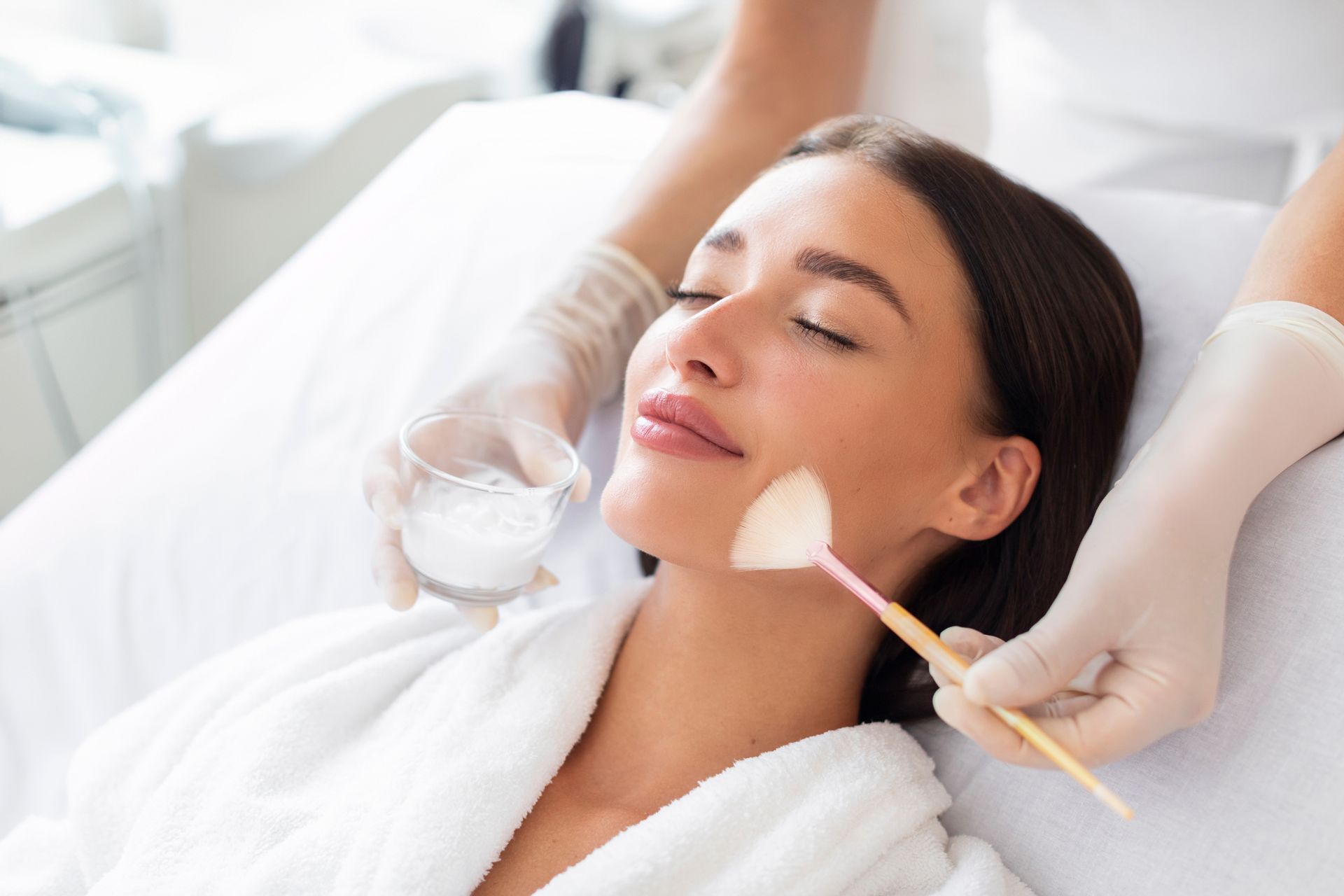 Person receiving a facial treatment while lying on a spa bed, with cream being applied by gloved hands