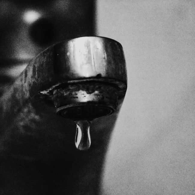 Close-up, black and white photo of a dripping faucet. Water droplet hangs beneath the spout.