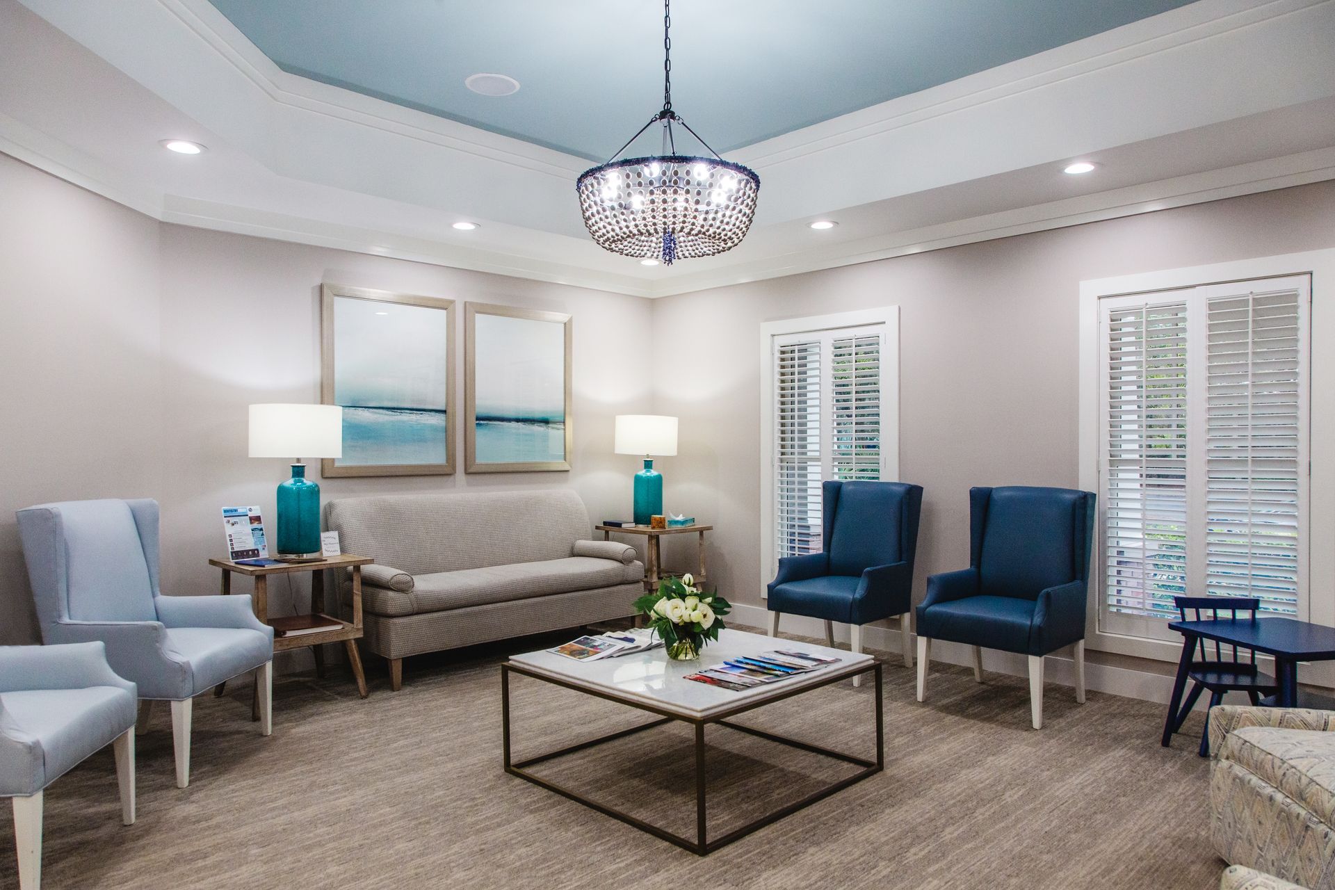 Bright modern sitting room with blue chairs, gray sofa, glass coffee table, lamps, and chandelier
