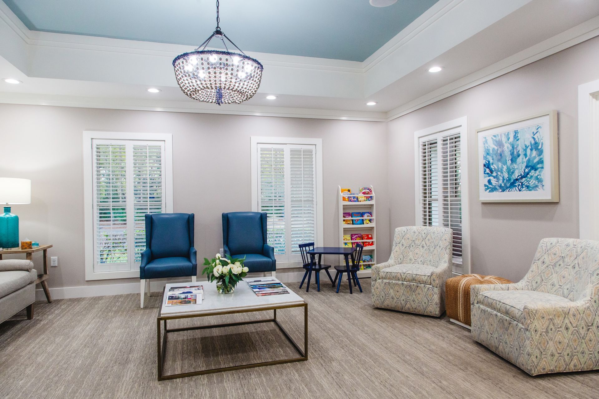 Bright living room with blue chairs, beige sofas, a coffee table, chandelier, and white shutters.